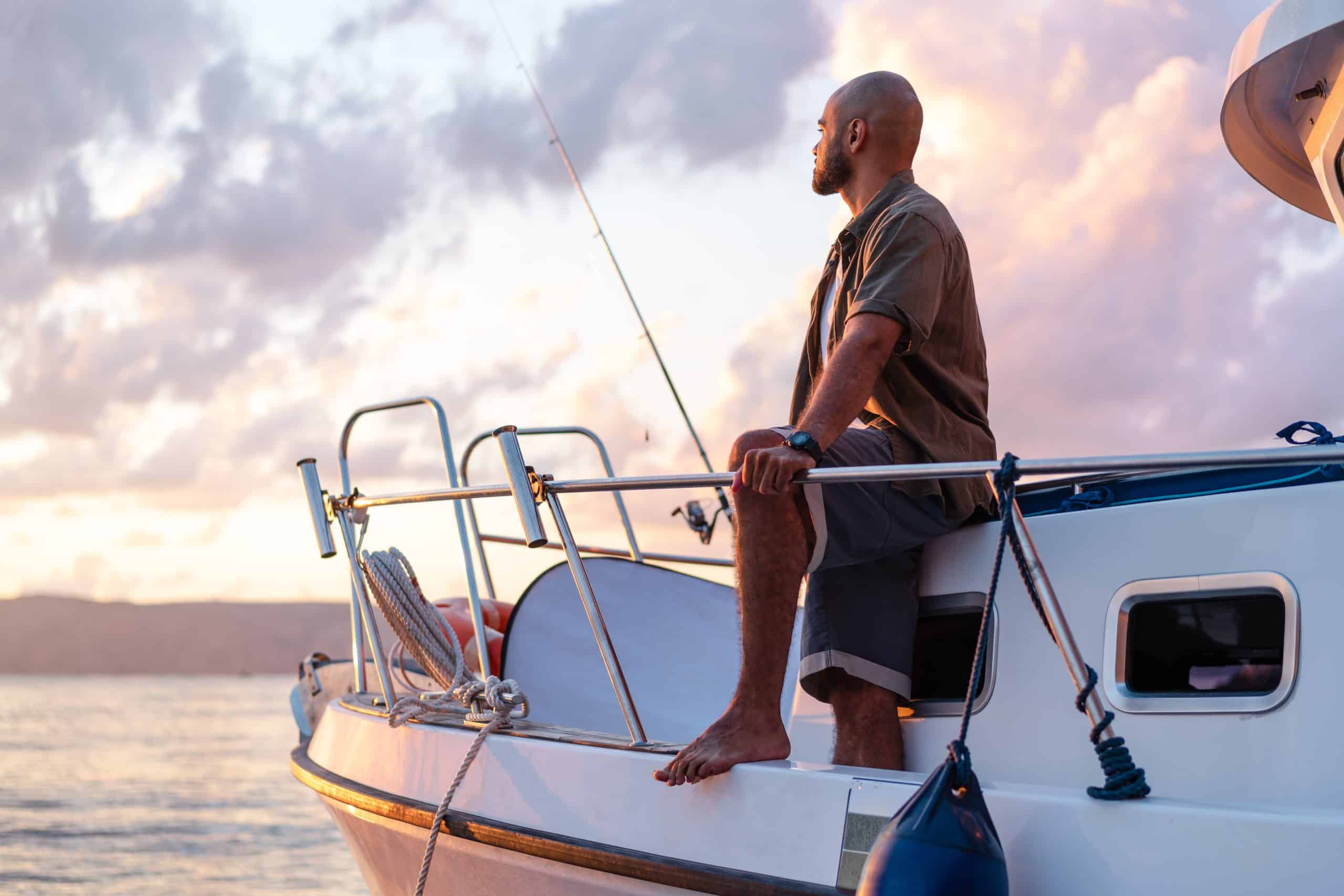 Young african american man standing with fishing rod on a sailboat fishing in open sea on sunset