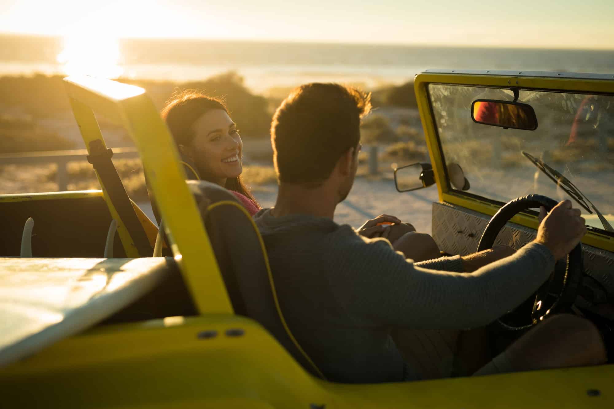 Happy caucasian couple sitting in beach buggy by the sea during sunset